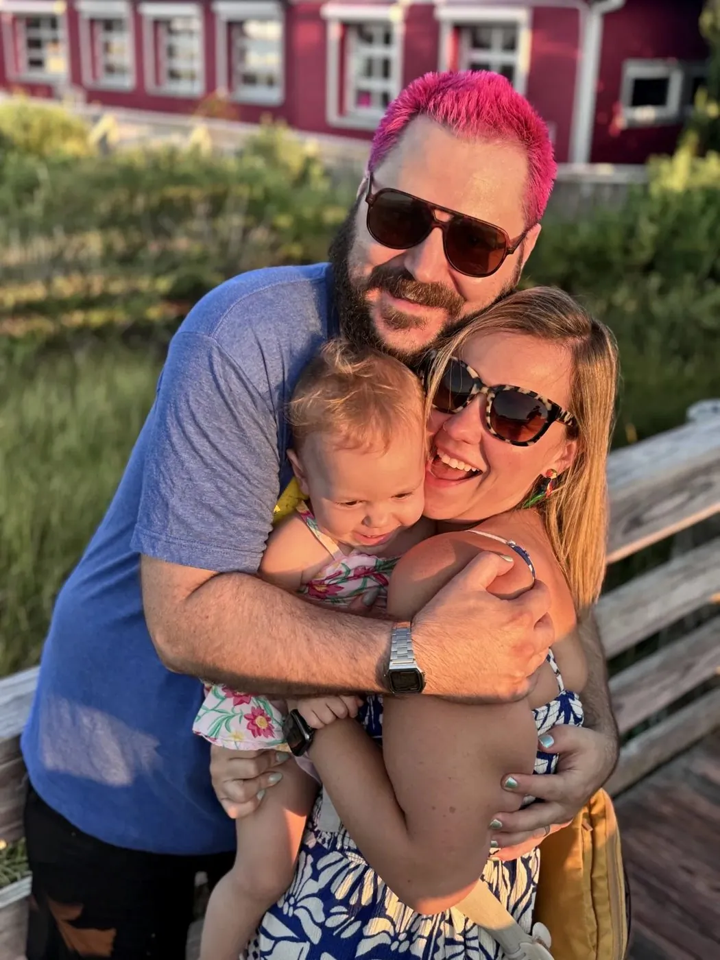 Adam, his wife and their toddler in a three-way hug on a sunny boardwalk