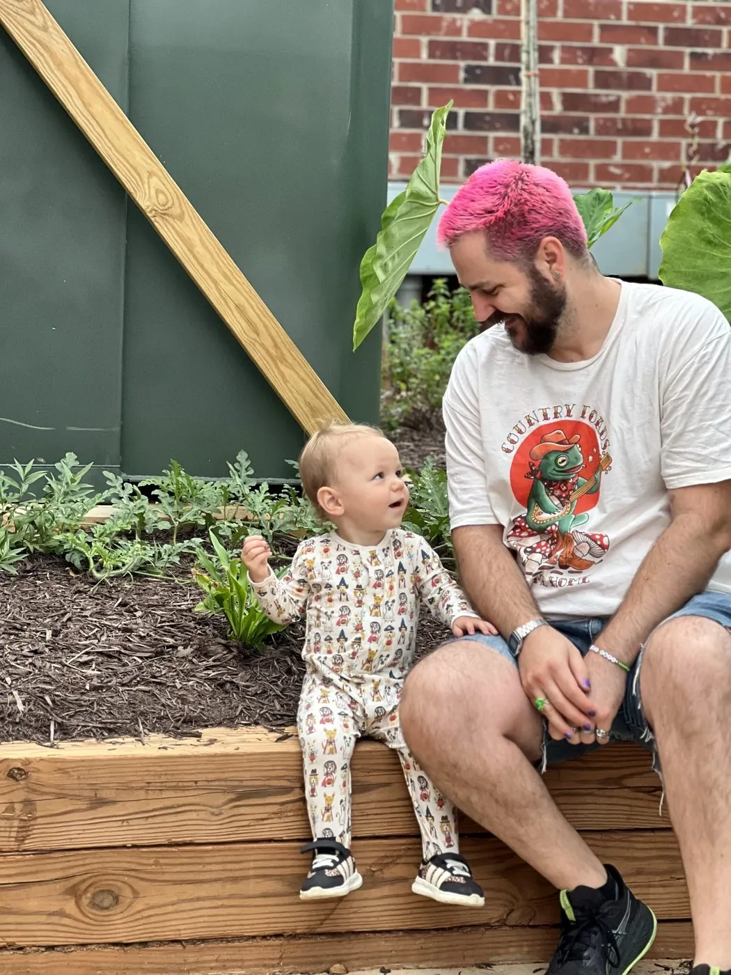 Adam with pink hair sitting on a garden bed with his baby, who grins up at him