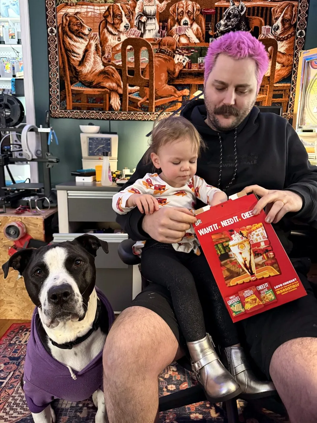 Adam at his home studio desk with his daughter on his lap and the dog in a purple hoodie beside them, under a dogs-playing-poker tapestry