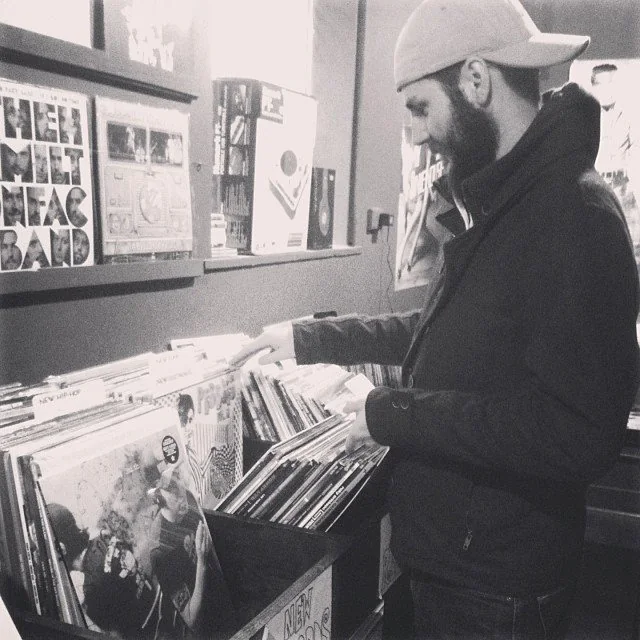 A black-and-white photo of Adam in a ball cap flipping through the new arrivals bins at a record store
